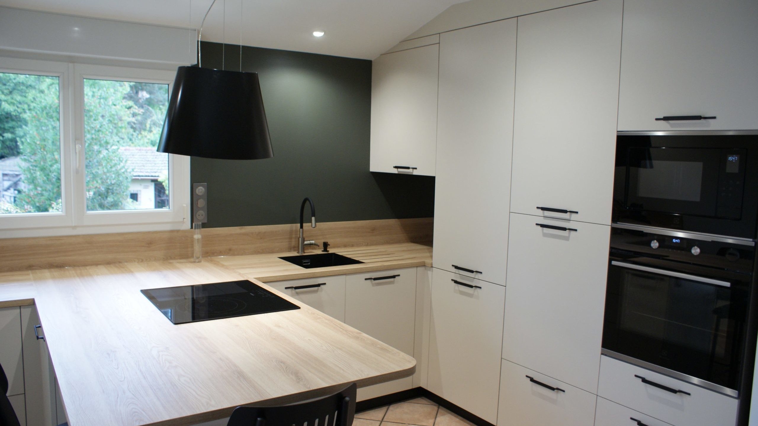 Cuisine moderne Miton Menta finition sabbia opaco, plans de travail et crédences en stratifié décor bois clair, îlot contre-mur avec plaque de cuisson, hotte suspendue Juno, four vapeur et lave-vaisselle intégré.