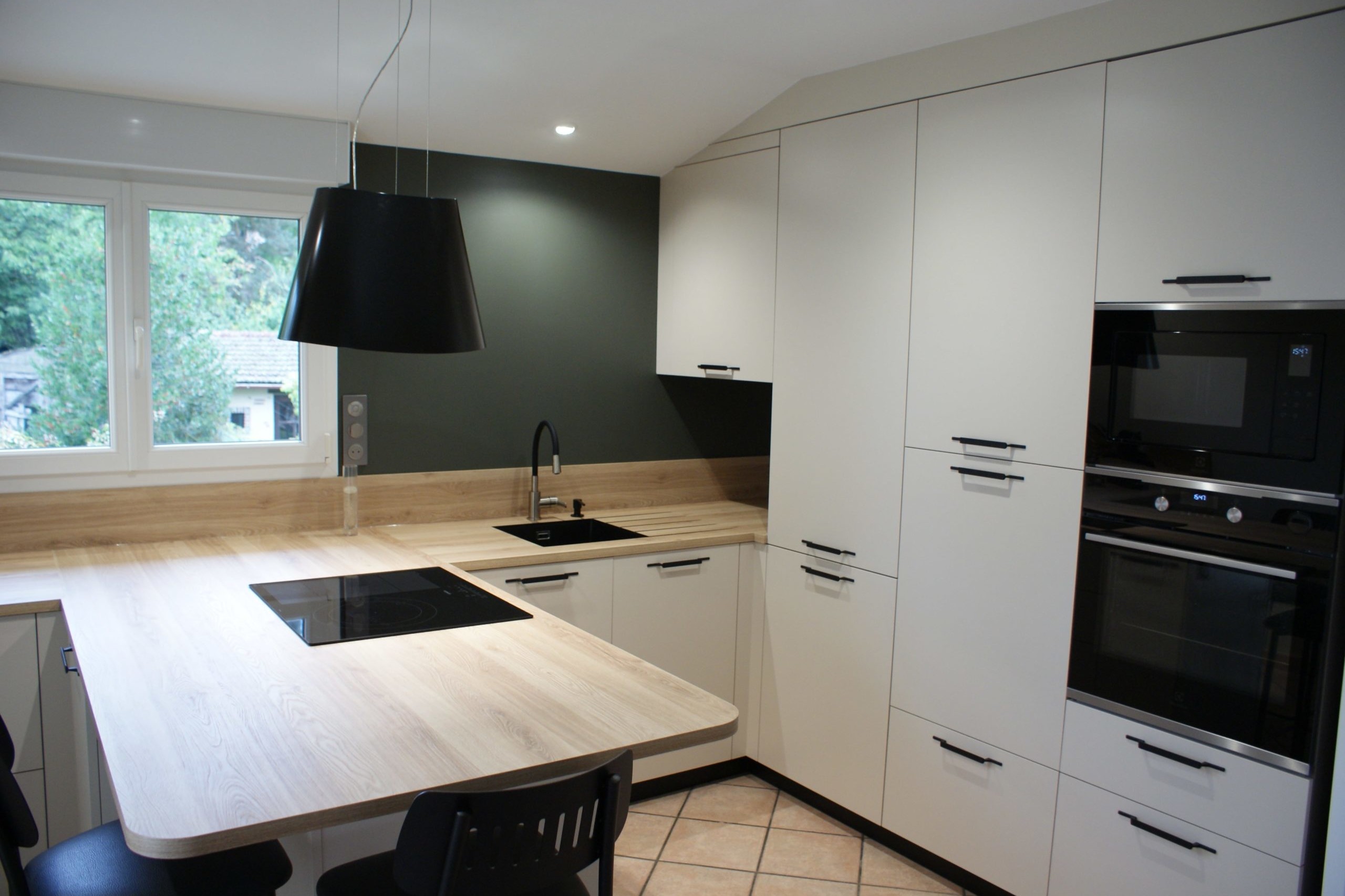 Cuisine moderne Miton Menta finition sabbia opaco, plans de travail et crédences en stratifié décor bois clair, îlot contre-mur avec plaque de cuisson, hotte suspendue Juno, four vapeur et lave-vaisselle intégré.