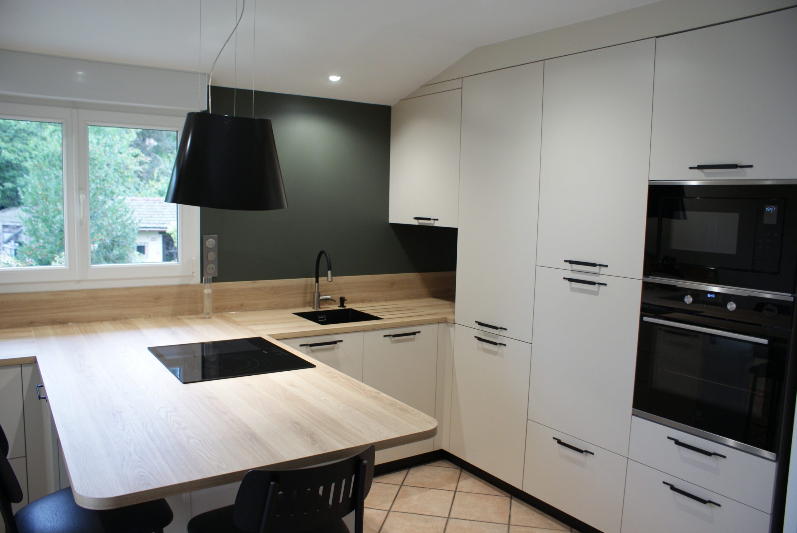 Cuisine moderne Miton Menta finition sabbia opaco, plans de travail et crédences en stratifié décor bois clair, îlot contre-mur avec plaque de cuisson, hotte suspendue Juno, four vapeur et lave-vaisselle intégré.