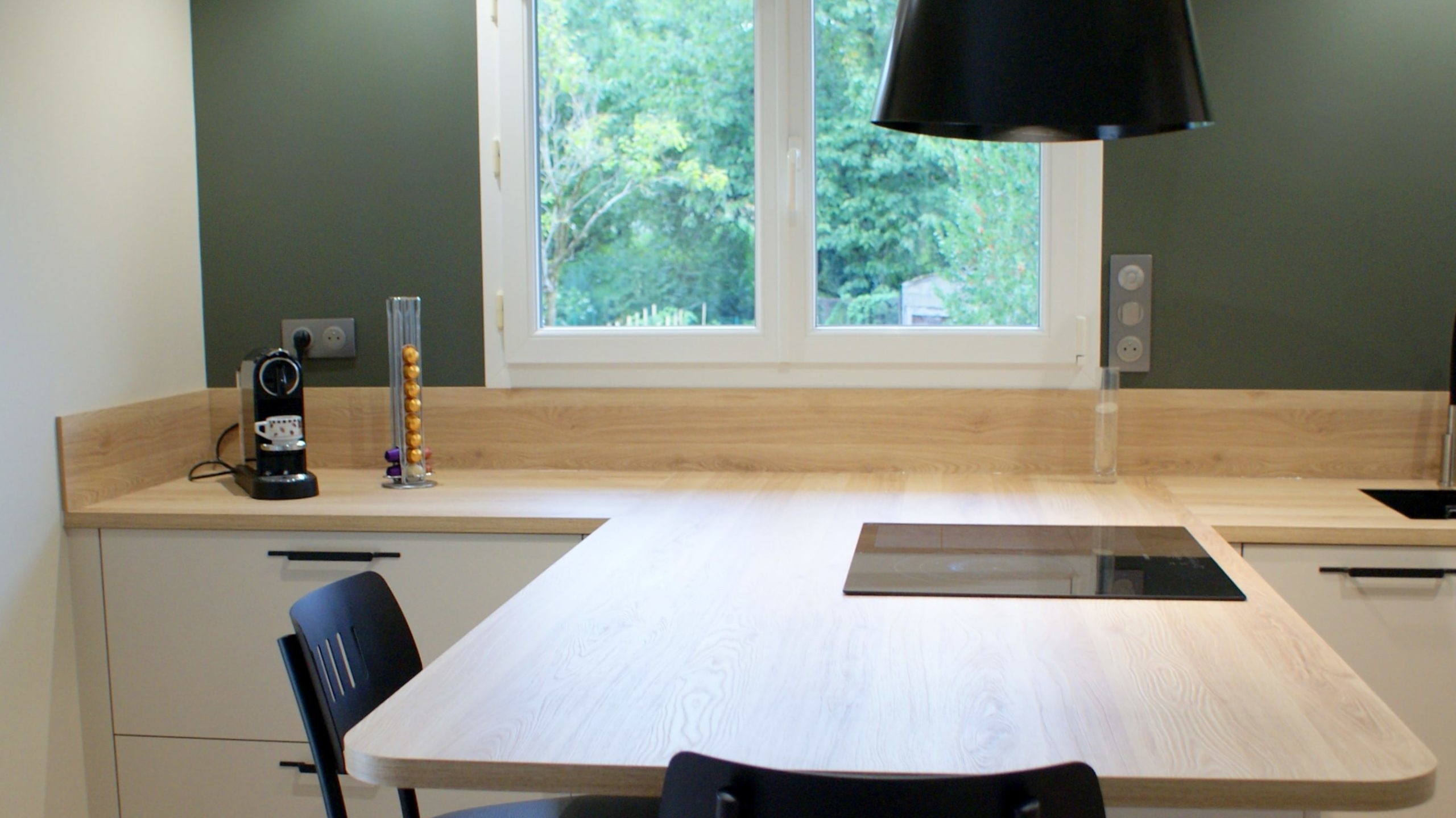 Cuisine moderne Miton Menta finition sabbia opaco, plans de travail et crédences en stratifié décor bois clair, îlot contre-mur avec plaque de cuisson, hotte suspendue Juno, four vapeur et lave-vaisselle intégré.