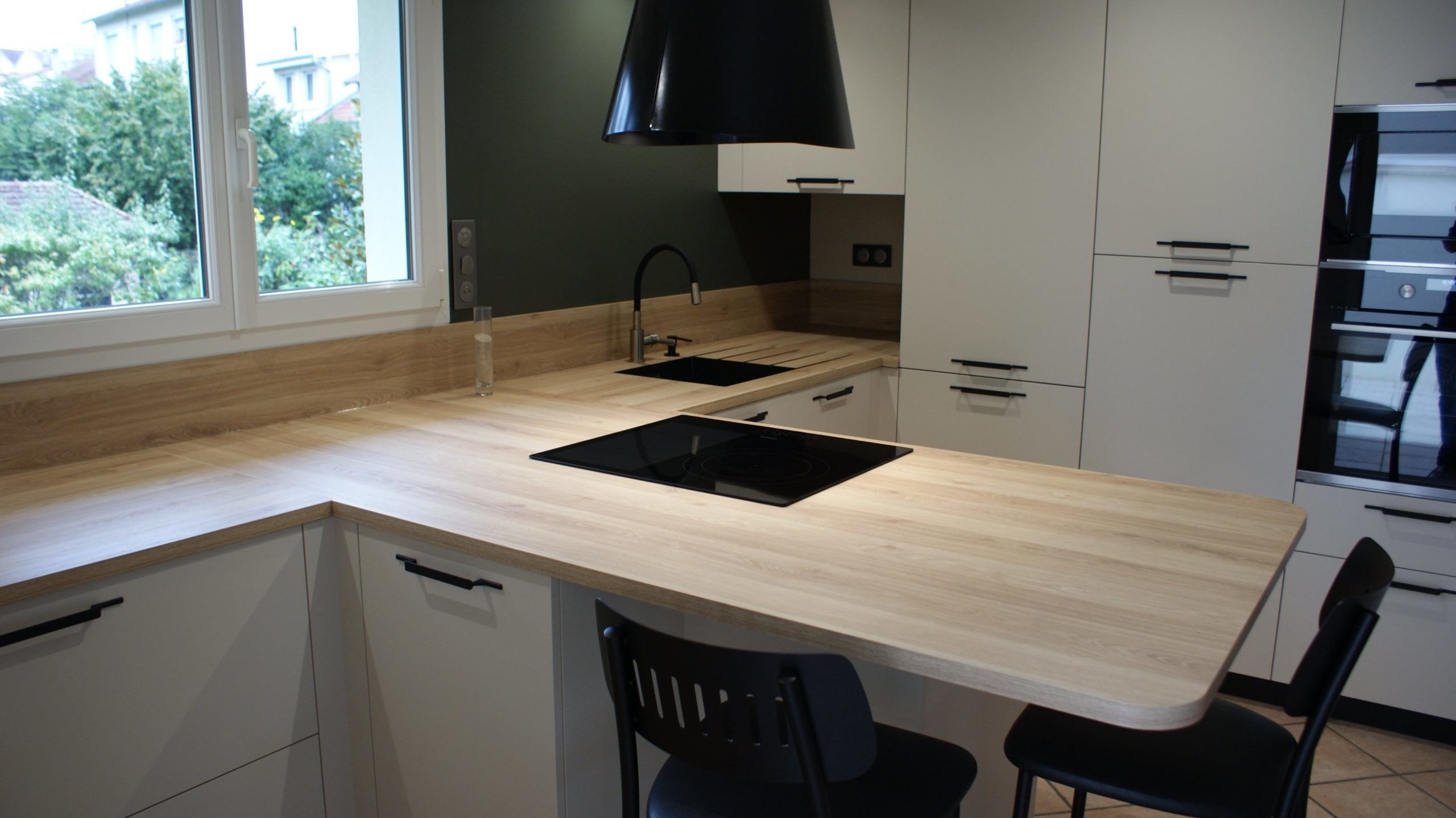 Cuisine moderne Miton Menta finition sabbia opaco, plans de travail et crédences en stratifié décor bois clair, îlot contre-mur avec plaque de cuisson, hotte suspendue Juno, four vapeur et lave-vaisselle intégré.