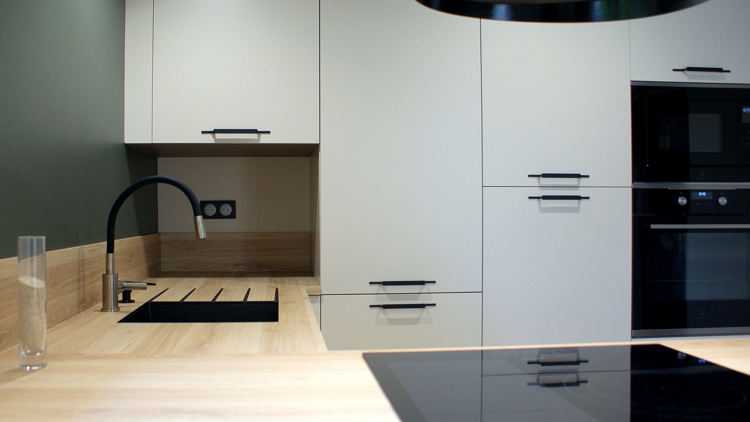 Cuisine moderne Miton Menta finition sabbia opaco, plans de travail et crédences en stratifié décor bois clair, îlot contre-mur avec plaque de cuisson, hotte suspendue Juno, four vapeur et lave-vaisselle intégré.