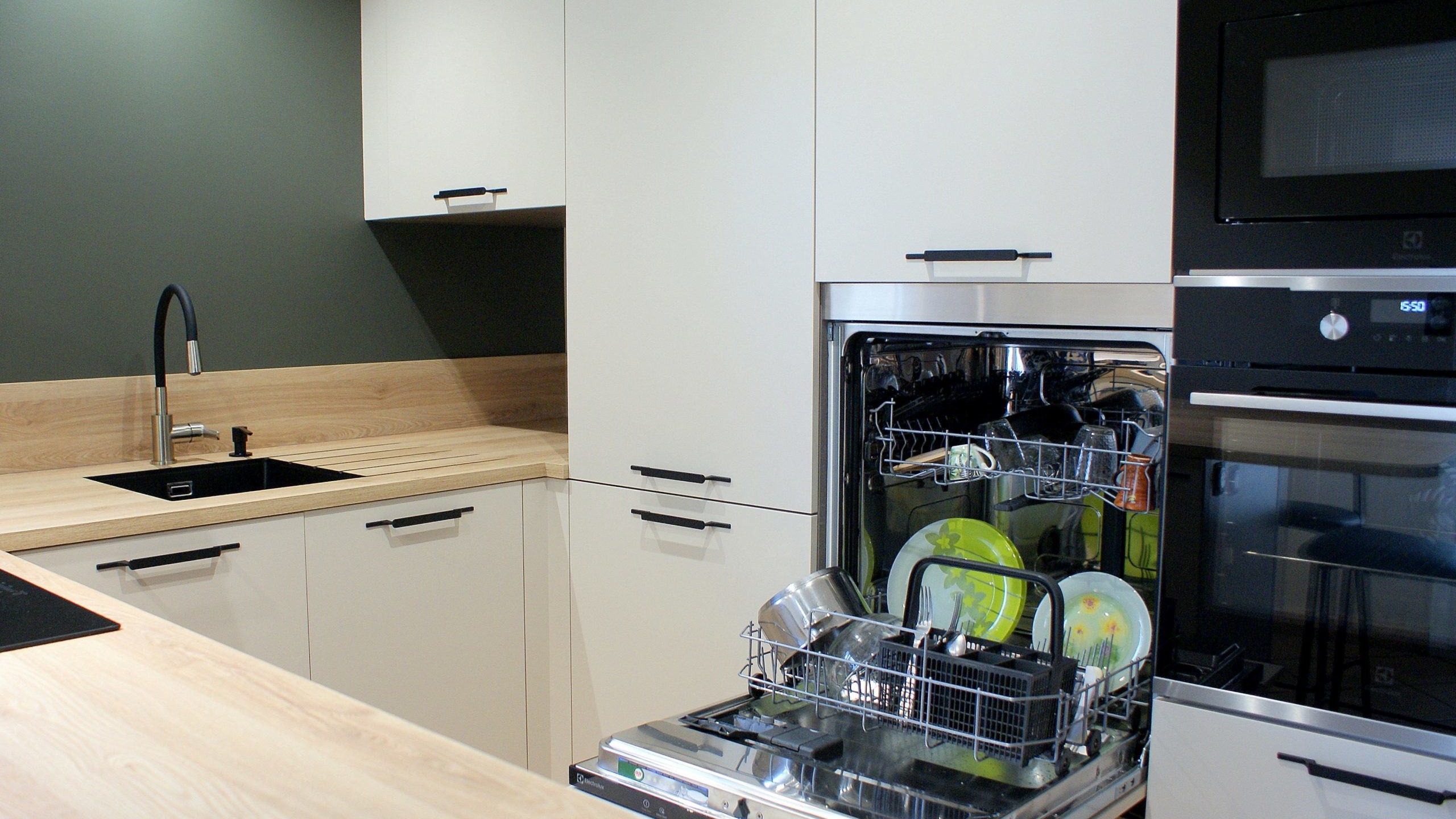 Cuisine moderne Miton Menta finition sabbia opaco, plans de travail et crédences en stratifié décor bois clair, îlot contre-mur avec plaque de cuisson, hotte suspendue Juno, four vapeur et lave-vaisselle intégré.