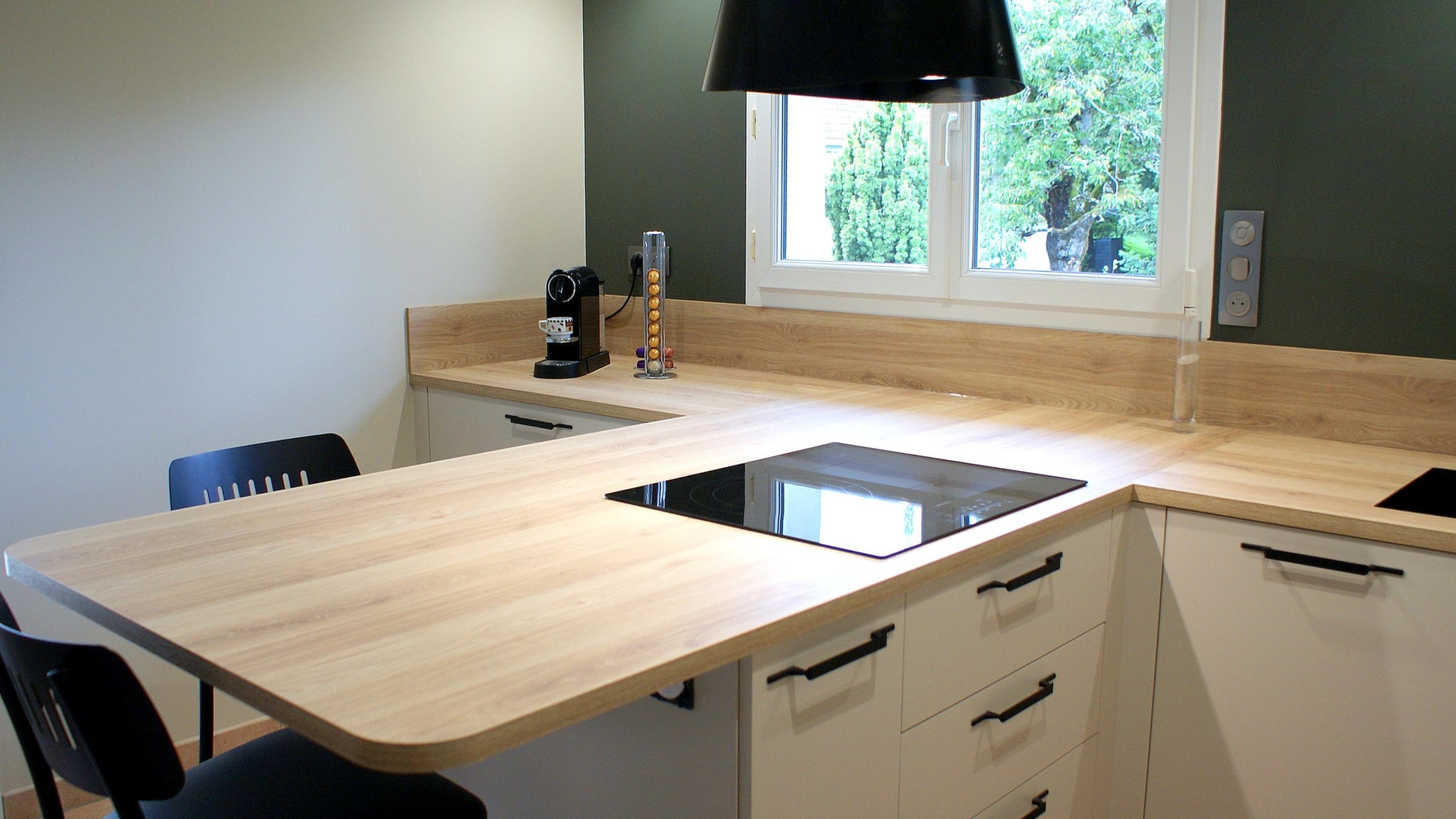 Cuisine moderne Miton Menta finition sabbia opaco, plans de travail et crédences en stratifié décor bois clair, îlot contre-mur avec plaque de cuisson, hotte suspendue Juno, four vapeur et lave-vaisselle intégré.