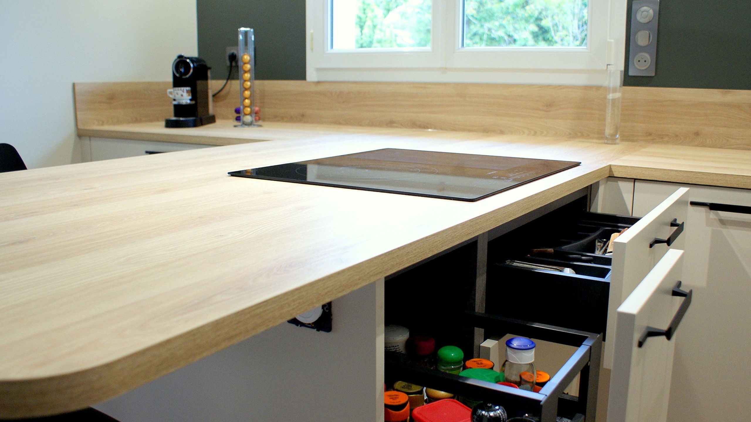 Cuisine moderne Miton Menta finition sabbia opaco, plans de travail et crédences en stratifié décor bois clair, îlot contre-mur avec plaque de cuisson, hotte suspendue Juno, four vapeur et lave-vaisselle intégré.