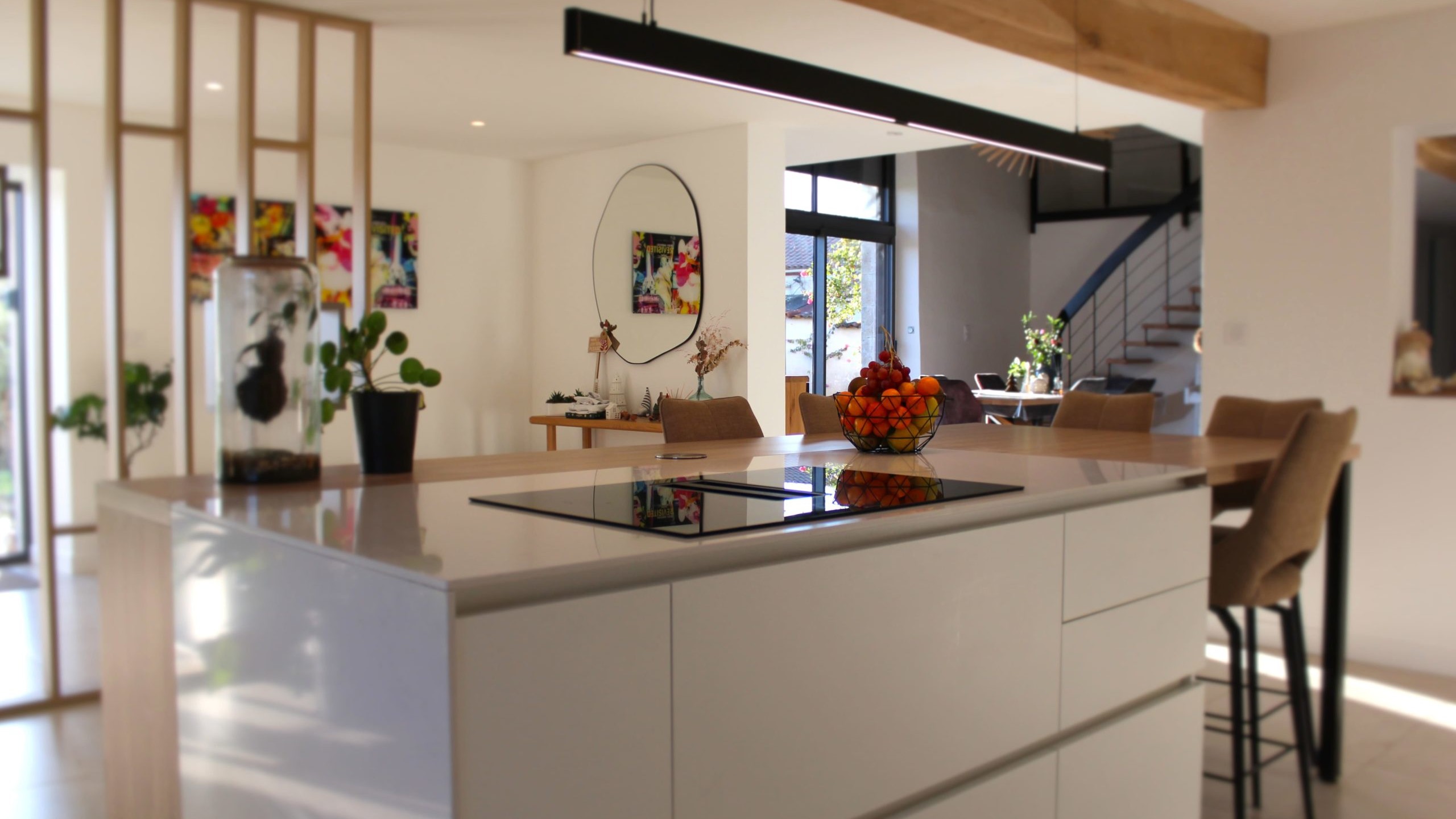 Cette cuisine contemporaine a été pensée comme un véritable espace de vie, alliant esthétique minimaliste, matériaux naturels et fonctionnalité au quotidien. L’association du bois clair et des façades blanches sans poignées apporte une sensation immédiate de luminosité et de sérénité. Au cœur de la pièce, l’îlot central s’impose comme un élément structurant. Il intègre une plaque de cuisson discrète, de nombreux rangements et une grande table en bois massif, idéale pour partager les repas, travailler ou recevoir. Cet aménagement favorise la convivialité tout en conservant une circulation fluide dans l’espace. La poutre en bois apparente traverse la pièce et lui confère un caractère unique, créant un dialogue harmonieux entre architecture traditionnelle et design contemporain. L’éclairage suspendu, aux lignes sobres et graphiques, souligne l’îlot et apporte une touche moderne élégante. Les espaces de rangement intégrés, l’électroménager encastré et les finitions soignées participent à une lecture visuelle épurée. Les touches végétales viennent parfaire l’ensemble en apportant fraîcheur et naturel. Pensée dans les moindres détails, cette cuisine offre un cadre à la fois chaleureux, fonctionnel et durable, parfaitement adapté à un art de vivre moderne.