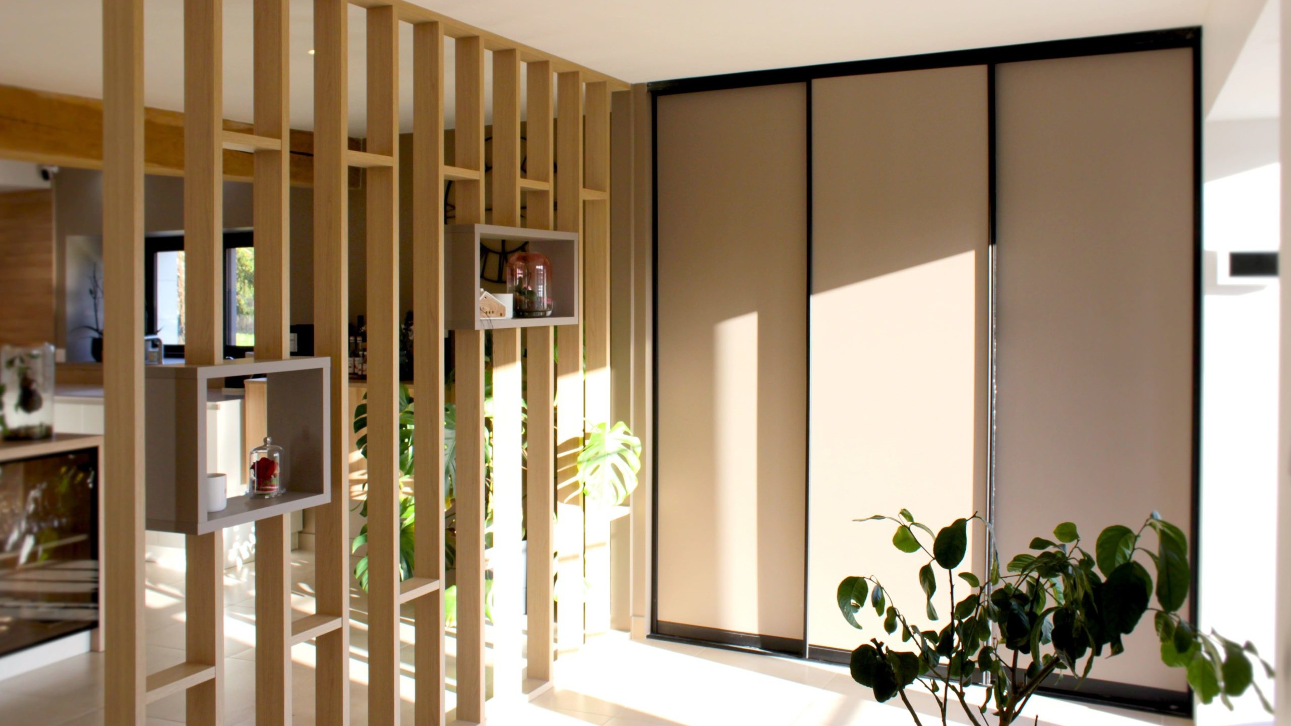 Cette cuisine contemporaine a été pensée comme un véritable espace de vie, alliant esthétique minimaliste, matériaux naturels et fonctionnalité au quotidien. L’association du bois clair et des façades blanches sans poignées apporte une sensation immédiate de luminosité et de sérénité. Au cœur de la pièce, l’îlot central s’impose comme un élément structurant. Il intègre une plaque de cuisson discrète, de nombreux rangements et une grande table en bois massif, idéale pour partager les repas, travailler ou recevoir. Cet aménagement favorise la convivialité tout en conservant une circulation fluide dans l’espace. La poutre en bois apparente traverse la pièce et lui confère un caractère unique, créant un dialogue harmonieux entre architecture traditionnelle et design contemporain. L’éclairage suspendu, aux lignes sobres et graphiques, souligne l’îlot et apporte une touche moderne élégante. Les espaces de rangement intégrés, l’électroménager encastré et les finitions soignées participent à une lecture visuelle épurée. Les touches végétales viennent parfaire l’ensemble en apportant fraîcheur et naturel. Pensée dans les moindres détails, cette cuisine offre un cadre à la fois chaleureux, fonctionnel et durable, parfaitement adapté à un art de vivre moderne.