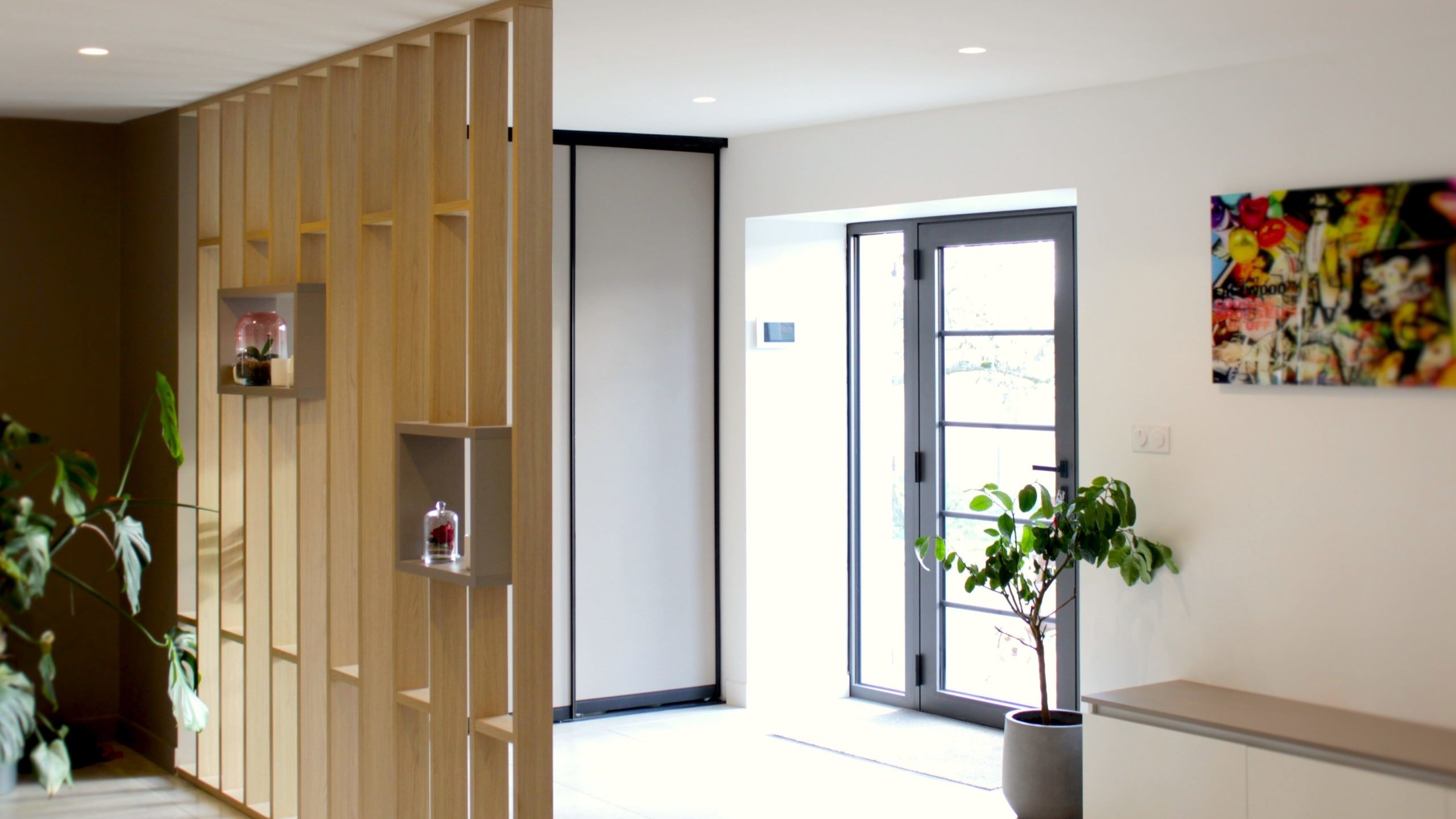 Cette cuisine contemporaine a été pensée comme un véritable espace de vie, alliant esthétique minimaliste, matériaux naturels et fonctionnalité au quotidien. L’association du bois clair et des façades blanches sans poignées apporte une sensation immédiate de luminosité et de sérénité. Au cœur de la pièce, l’îlot central s’impose comme un élément structurant. Il intègre une plaque de cuisson discrète, de nombreux rangements et une grande table en bois massif, idéale pour partager les repas, travailler ou recevoir. Cet aménagement favorise la convivialité tout en conservant une circulation fluide dans l’espace. La poutre en bois apparente traverse la pièce et lui confère un caractère unique, créant un dialogue harmonieux entre architecture traditionnelle et design contemporain. L’éclairage suspendu, aux lignes sobres et graphiques, souligne l’îlot et apporte une touche moderne élégante. Les espaces de rangement intégrés, l’électroménager encastré et les finitions soignées participent à une lecture visuelle épurée. Les touches végétales viennent parfaire l’ensemble en apportant fraîcheur et naturel. Pensée dans les moindres détails, cette cuisine offre un cadre à la fois chaleureux, fonctionnel et durable, parfaitement adapté à un art de vivre moderne.