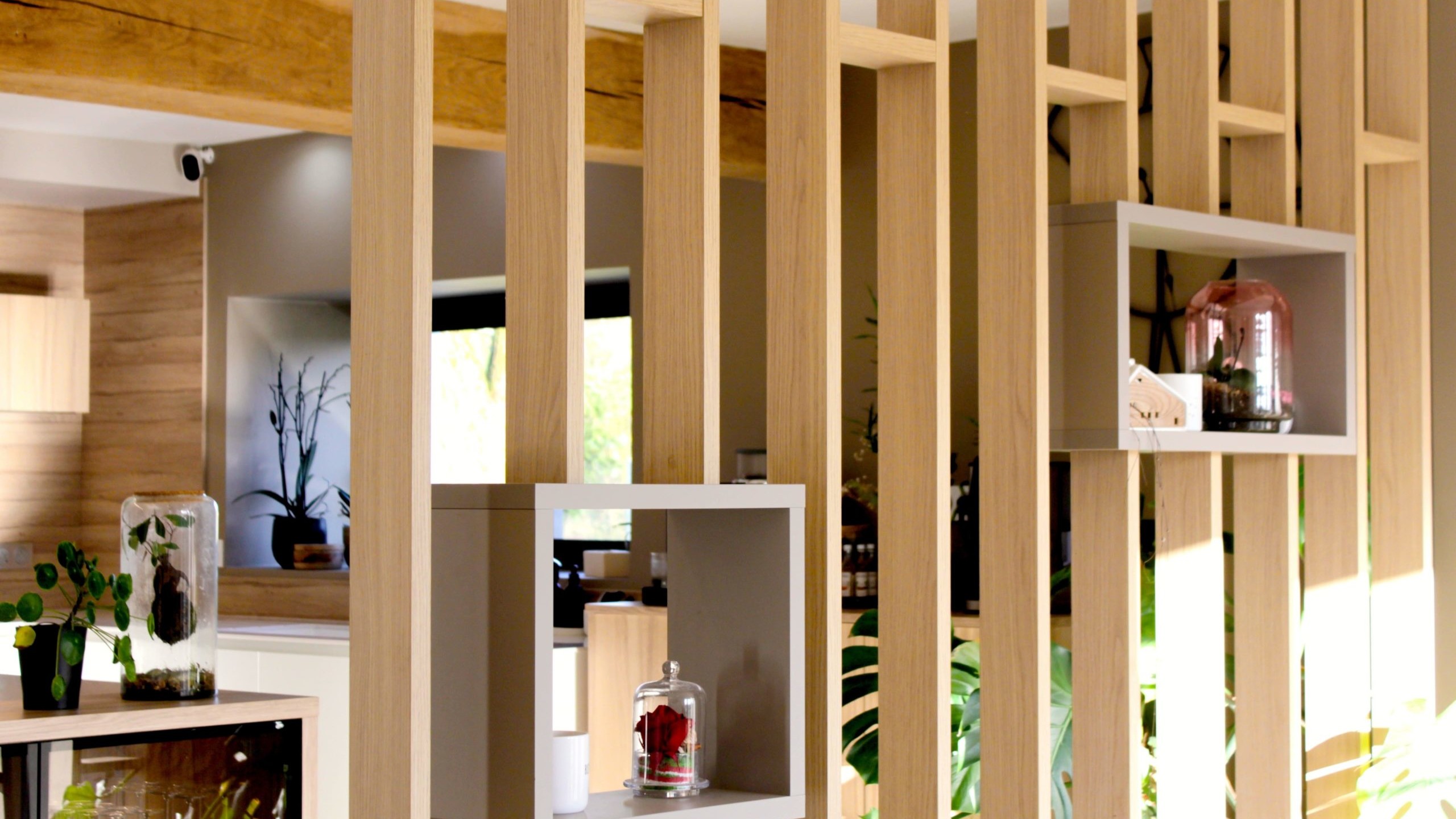 Cette cuisine contemporaine a été pensée comme un véritable espace de vie, alliant esthétique minimaliste, matériaux naturels et fonctionnalité au quotidien. L’association du bois clair et des façades blanches sans poignées apporte une sensation immédiate de luminosité et de sérénité. Au cœur de la pièce, l’îlot central s’impose comme un élément structurant. Il intègre une plaque de cuisson discrète, de nombreux rangements et une grande table en bois massif, idéale pour partager les repas, travailler ou recevoir. Cet aménagement favorise la convivialité tout en conservant une circulation fluide dans l’espace. La poutre en bois apparente traverse la pièce et lui confère un caractère unique, créant un dialogue harmonieux entre architecture traditionnelle et design contemporain. L’éclairage suspendu, aux lignes sobres et graphiques, souligne l’îlot et apporte une touche moderne élégante. Les espaces de rangement intégrés, l’électroménager encastré et les finitions soignées participent à une lecture visuelle épurée. Les touches végétales viennent parfaire l’ensemble en apportant fraîcheur et naturel. Pensée dans les moindres détails, cette cuisine offre un cadre à la fois chaleureux, fonctionnel et durable, parfaitement adapté à un art de vivre moderne.