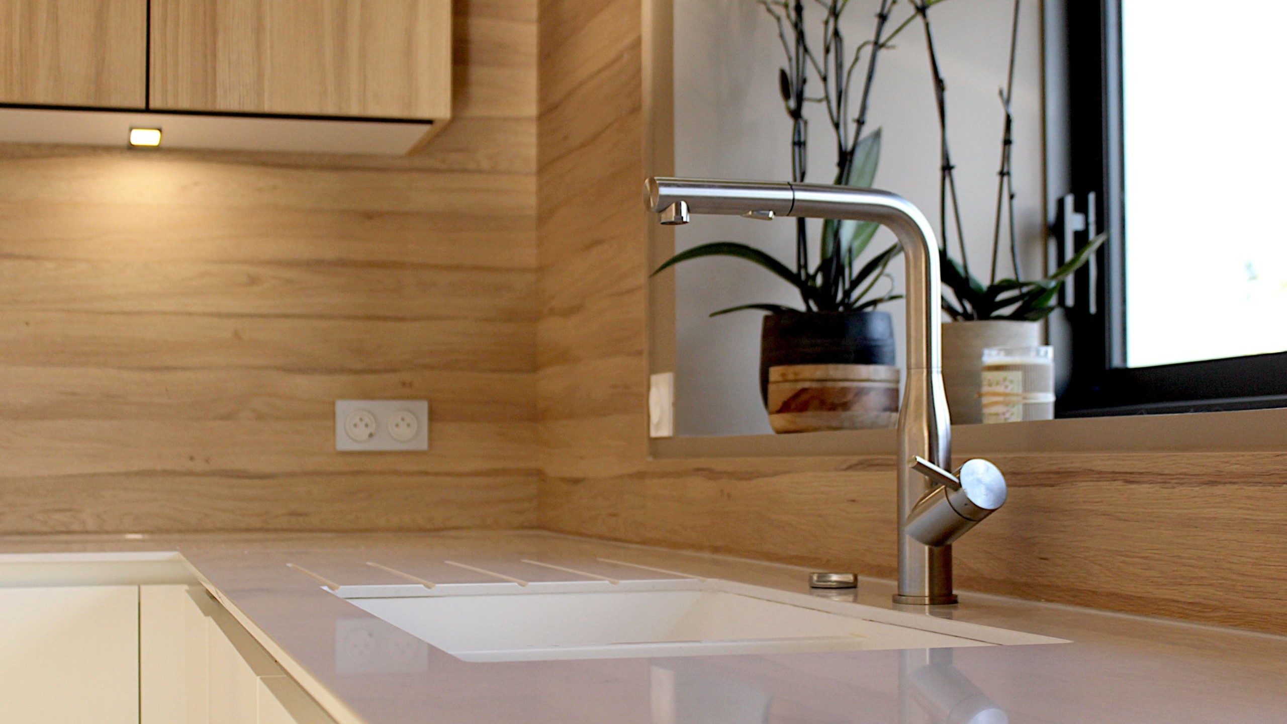 Cette cuisine contemporaine a été pensée comme un véritable espace de vie, alliant esthétique minimaliste, matériaux naturels et fonctionnalité au quotidien. L’association du bois clair et des façades blanches sans poignées apporte une sensation immédiate de luminosité et de sérénité. Au cœur de la pièce, l’îlot central s’impose comme un élément structurant. Il intègre une plaque de cuisson discrète, de nombreux rangements et une grande table en bois massif, idéale pour partager les repas, travailler ou recevoir. Cet aménagement favorise la convivialité tout en conservant une circulation fluide dans l’espace. La poutre en bois apparente traverse la pièce et lui confère un caractère unique, créant un dialogue harmonieux entre architecture traditionnelle et design contemporain. L’éclairage suspendu, aux lignes sobres et graphiques, souligne l’îlot et apporte une touche moderne élégante. Les espaces de rangement intégrés, l’électroménager encastré et les finitions soignées participent à une lecture visuelle épurée. Les touches végétales viennent parfaire l’ensemble en apportant fraîcheur et naturel. Pensée dans les moindres détails, cette cuisine offre un cadre à la fois chaleureux, fonctionnel et durable, parfaitement adapté à un art de vivre moderne.