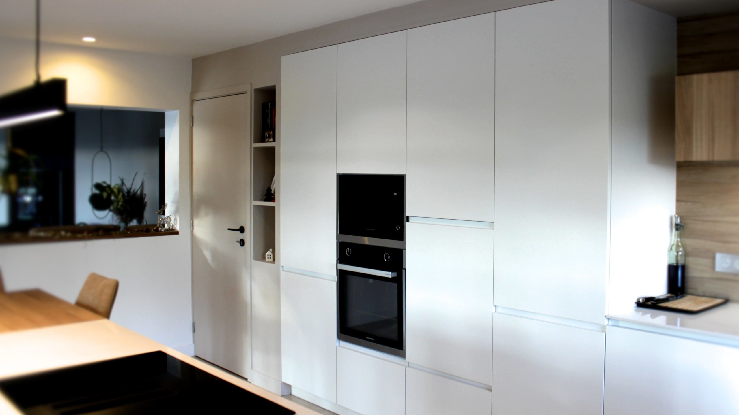 Cette cuisine contemporaine a été pensée comme un véritable espace de vie, alliant esthétique minimaliste, matériaux naturels et fonctionnalité au quotidien. L’association du bois clair et des façades blanches sans poignées apporte une sensation immédiate de luminosité et de sérénité. Au cœur de la pièce, l’îlot central s’impose comme un élément structurant. Il intègre une plaque de cuisson discrète, de nombreux rangements et une grande table en bois massif, idéale pour partager les repas, travailler ou recevoir. Cet aménagement favorise la convivialité tout en conservant une circulation fluide dans l’espace. La poutre en bois apparente traverse la pièce et lui confère un caractère unique, créant un dialogue harmonieux entre architecture traditionnelle et design contemporain. L’éclairage suspendu, aux lignes sobres et graphiques, souligne l’îlot et apporte une touche moderne élégante. Les espaces de rangement intégrés, l’électroménager encastré et les finitions soignées participent à une lecture visuelle épurée. Les touches végétales viennent parfaire l’ensemble en apportant fraîcheur et naturel. Pensée dans les moindres détails, cette cuisine offre un cadre à la fois chaleureux, fonctionnel et durable, parfaitement adapté à un art de vivre moderne.