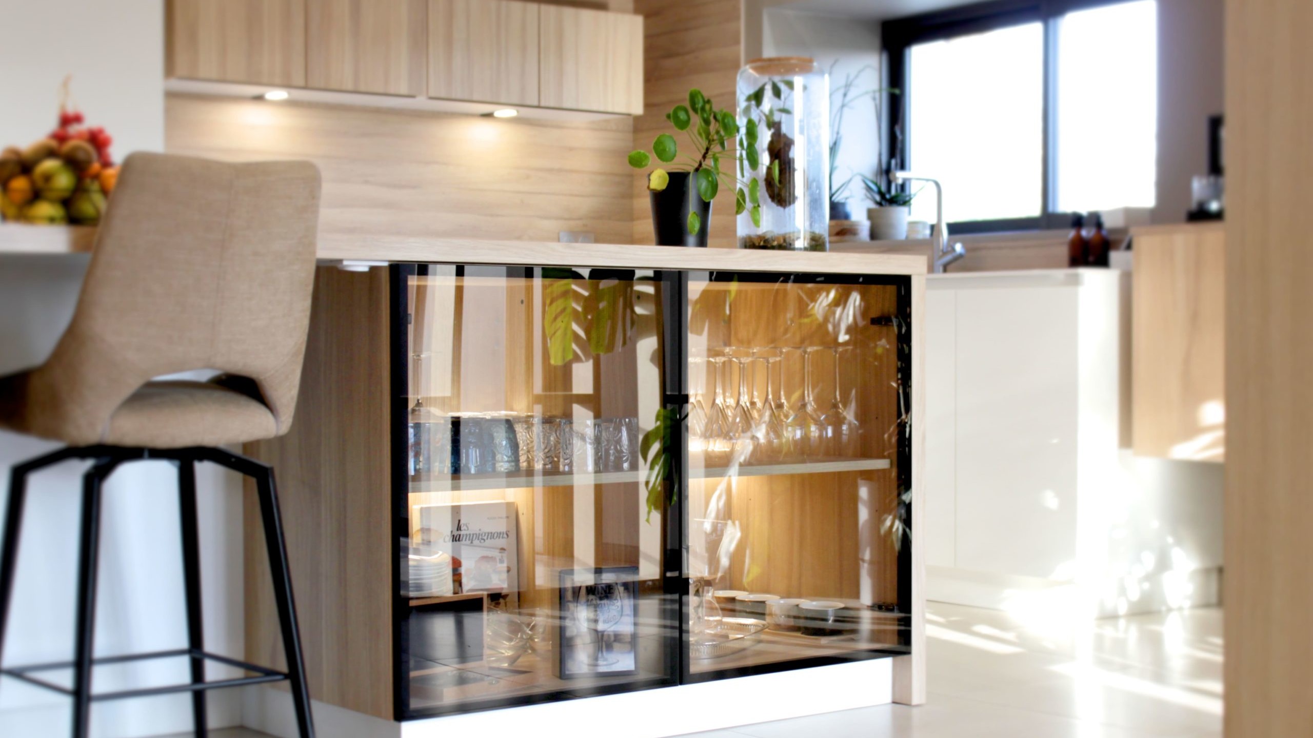 Cette cuisine contemporaine a été pensée comme un véritable espace de vie, alliant esthétique minimaliste, matériaux naturels et fonctionnalité au quotidien. L’association du bois clair et des façades blanches sans poignées apporte une sensation immédiate de luminosité et de sérénité. Au cœur de la pièce, l’îlot central s’impose comme un élément structurant. Il intègre une plaque de cuisson discrète, de nombreux rangements et une grande table en bois massif, idéale pour partager les repas, travailler ou recevoir. Cet aménagement favorise la convivialité tout en conservant une circulation fluide dans l’espace. La poutre en bois apparente traverse la pièce et lui confère un caractère unique, créant un dialogue harmonieux entre architecture traditionnelle et design contemporain. L’éclairage suspendu, aux lignes sobres et graphiques, souligne l’îlot et apporte une touche moderne élégante. Les espaces de rangement intégrés, l’électroménager encastré et les finitions soignées participent à une lecture visuelle épurée. Les touches végétales viennent parfaire l’ensemble en apportant fraîcheur et naturel. Pensée dans les moindres détails, cette cuisine offre un cadre à la fois chaleureux, fonctionnel et durable, parfaitement adapté à un art de vivre moderne.