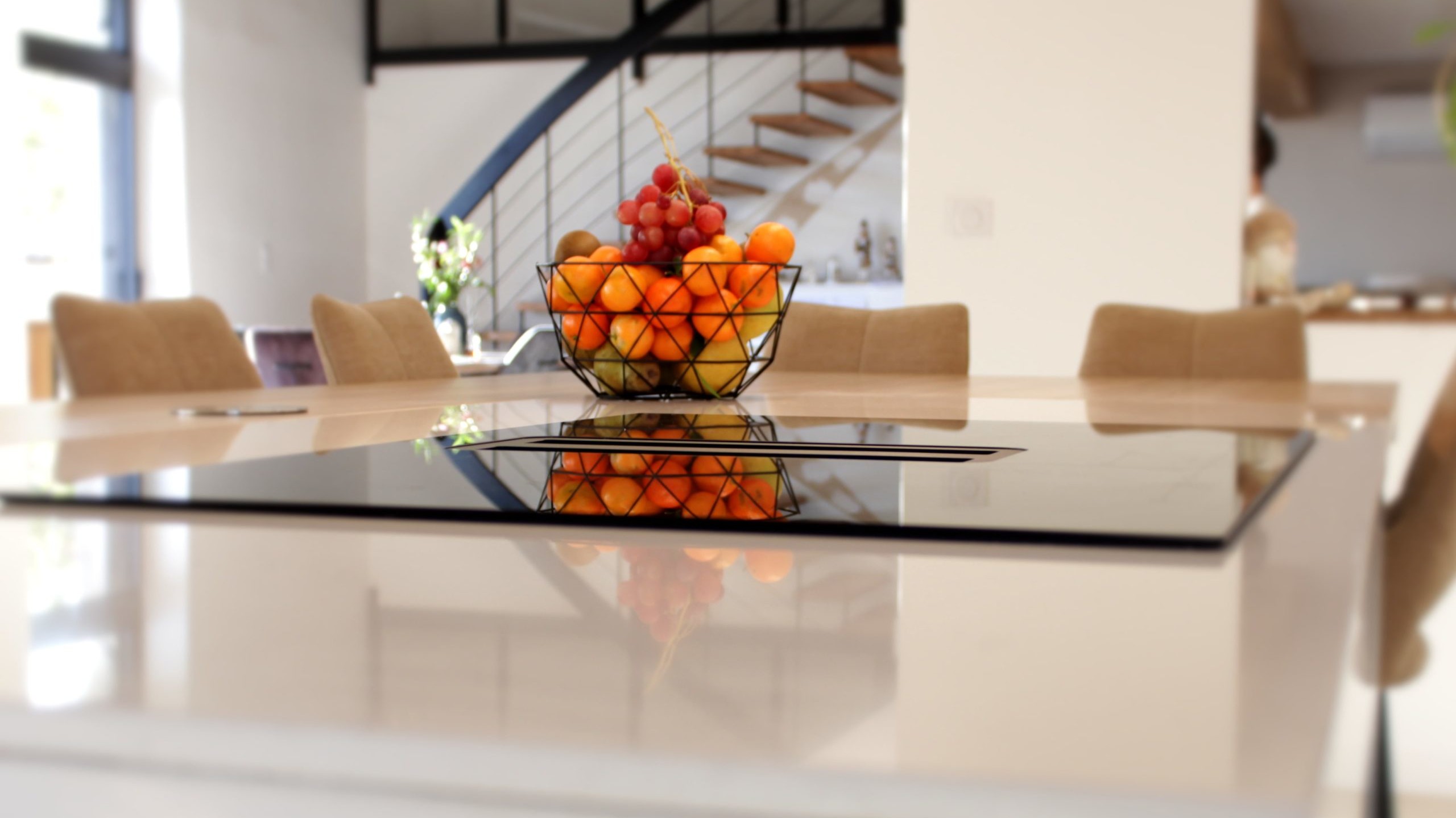 Cette cuisine contemporaine a été pensée comme un véritable espace de vie, alliant esthétique minimaliste, matériaux naturels et fonctionnalité au quotidien. L’association du bois clair et des façades blanches sans poignées apporte une sensation immédiate de luminosité et de sérénité. Au cœur de la pièce, l’îlot central s’impose comme un élément structurant. Il intègre une plaque de cuisson discrète, de nombreux rangements et une grande table en bois massif, idéale pour partager les repas, travailler ou recevoir. Cet aménagement favorise la convivialité tout en conservant une circulation fluide dans l’espace. La poutre en bois apparente traverse la pièce et lui confère un caractère unique, créant un dialogue harmonieux entre architecture traditionnelle et design contemporain. L’éclairage suspendu, aux lignes sobres et graphiques, souligne l’îlot et apporte une touche moderne élégante. Les espaces de rangement intégrés, l’électroménager encastré et les finitions soignées participent à une lecture visuelle épurée. Les touches végétales viennent parfaire l’ensemble en apportant fraîcheur et naturel. Pensée dans les moindres détails, cette cuisine offre un cadre à la fois chaleureux, fonctionnel et durable, parfaitement adapté à un art de vivre moderne.