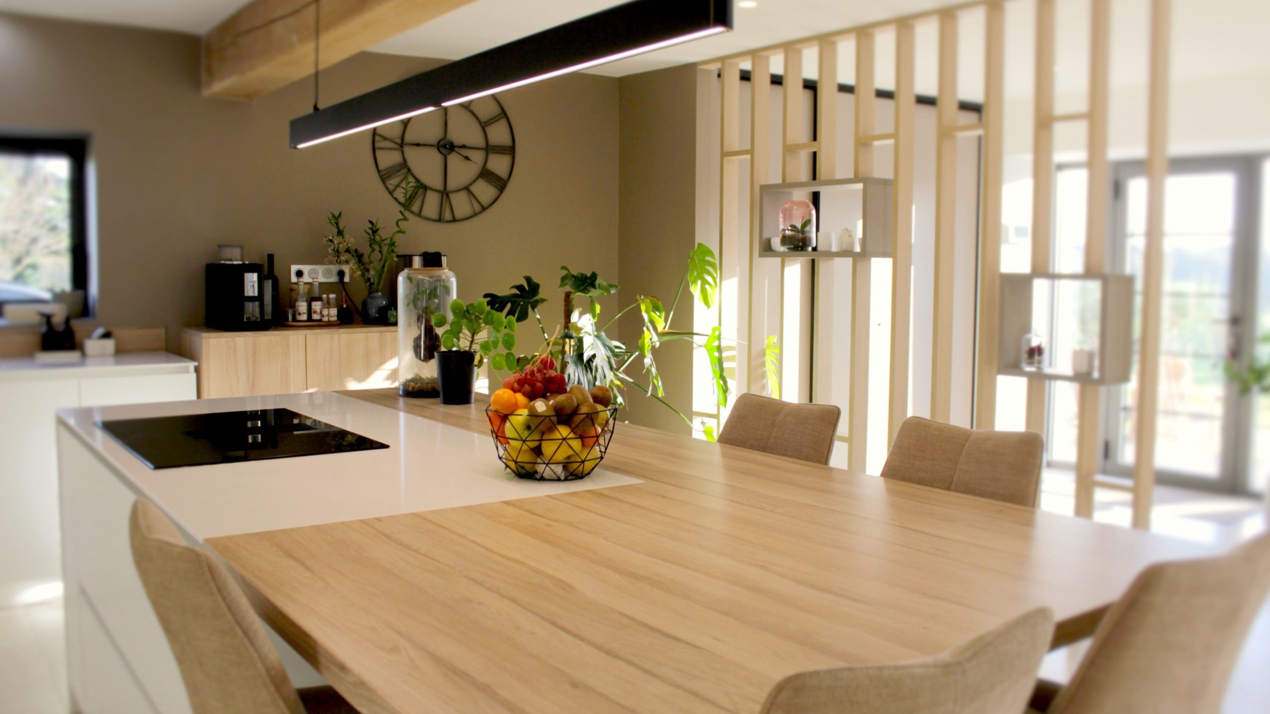 Cette cuisine contemporaine a été pensée comme un véritable espace de vie, alliant esthétique minimaliste, matériaux naturels et fonctionnalité au quotidien. L’association du bois clair et des façades blanches sans poignées apporte une sensation immédiate de luminosité et de sérénité. Au cœur de la pièce, l’îlot central s’impose comme un élément structurant. Il intègre une plaque de cuisson discrète, de nombreux rangements et une grande table en bois massif, idéale pour partager les repas, travailler ou recevoir. Cet aménagement favorise la convivialité tout en conservant une circulation fluide dans l’espace. La poutre en bois apparente traverse la pièce et lui confère un caractère unique, créant un dialogue harmonieux entre architecture traditionnelle et design contemporain. L’éclairage suspendu, aux lignes sobres et graphiques, souligne l’îlot et apporte une touche moderne élégante. Les espaces de rangement intégrés, l’électroménager encastré et les finitions soignées participent à une lecture visuelle épurée. Les touches végétales viennent parfaire l’ensemble en apportant fraîcheur et naturel. Pensée dans les moindres détails, cette cuisine offre un cadre à la fois chaleureux, fonctionnel et durable, parfaitement adapté à un art de vivre moderne.