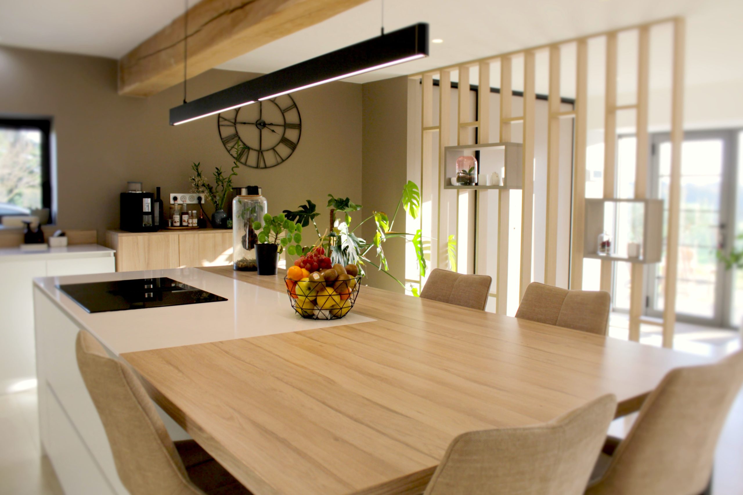 Cette cuisine contemporaine a été pensée comme un véritable espace de vie, alliant esthétique minimaliste, matériaux naturels et fonctionnalité au quotidien. L’association du bois clair et des façades blanches sans poignées apporte une sensation immédiate de luminosité et de sérénité. Au cœur de la pièce, l’îlot central s’impose comme un élément structurant. Il intègre une plaque de cuisson discrète, de nombreux rangements et une grande table en bois massif, idéale pour partager les repas, travailler ou recevoir. Cet aménagement favorise la convivialité tout en conservant une circulation fluide dans l’espace. La poutre en bois apparente traverse la pièce et lui confère un caractère unique, créant un dialogue harmonieux entre architecture traditionnelle et design contemporain. L’éclairage suspendu, aux lignes sobres et graphiques, souligne l’îlot et apporte une touche moderne élégante. Les espaces de rangement intégrés, l’électroménager encastré et les finitions soignées participent à une lecture visuelle épurée. Les touches végétales viennent parfaire l’ensemble en apportant fraîcheur et naturel. Pensée dans les moindres détails, cette cuisine offre un cadre à la fois chaleureux, fonctionnel et durable, parfaitement adapté à un art de vivre moderne.