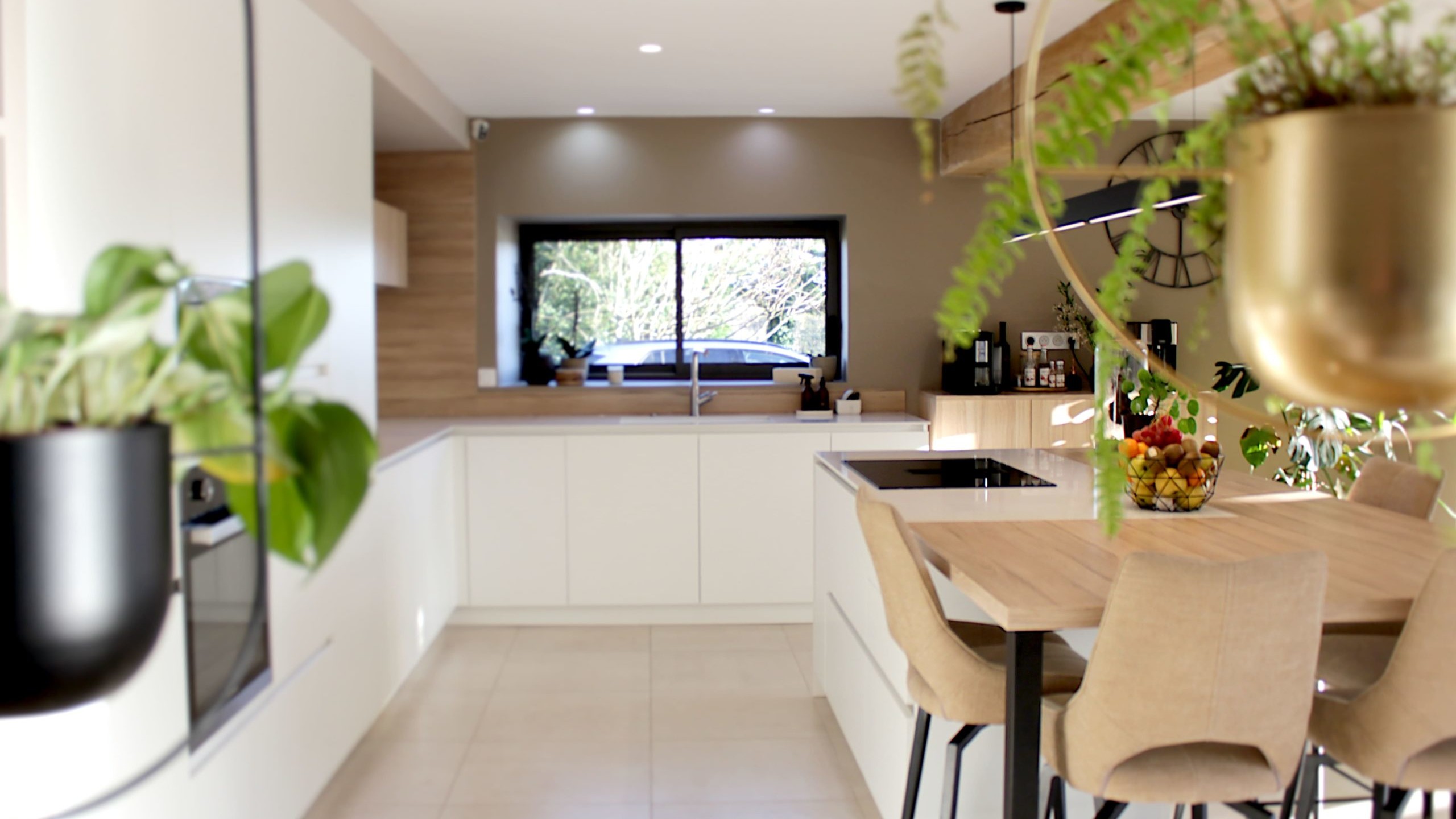 Cette cuisine contemporaine a été pensée comme un véritable espace de vie, alliant esthétique minimaliste, matériaux naturels et fonctionnalité au quotidien. L’association du bois clair et des façades blanches sans poignées apporte une sensation immédiate de luminosité et de sérénité. Au cœur de la pièce, l’îlot central s’impose comme un élément structurant. Il intègre une plaque de cuisson discrète, de nombreux rangements et une grande table en bois massif, idéale pour partager les repas, travailler ou recevoir. Cet aménagement favorise la convivialité tout en conservant une circulation fluide dans l’espace. La poutre en bois apparente traverse la pièce et lui confère un caractère unique, créant un dialogue harmonieux entre architecture traditionnelle et design contemporain. L’éclairage suspendu, aux lignes sobres et graphiques, souligne l’îlot et apporte une touche moderne élégante. Les espaces de rangement intégrés, l’électroménager encastré et les finitions soignées participent à une lecture visuelle épurée. Les touches végétales viennent parfaire l’ensemble en apportant fraîcheur et naturel. Pensée dans les moindres détails, cette cuisine offre un cadre à la fois chaleureux, fonctionnel et durable, parfaitement adapté à un art de vivre moderne.