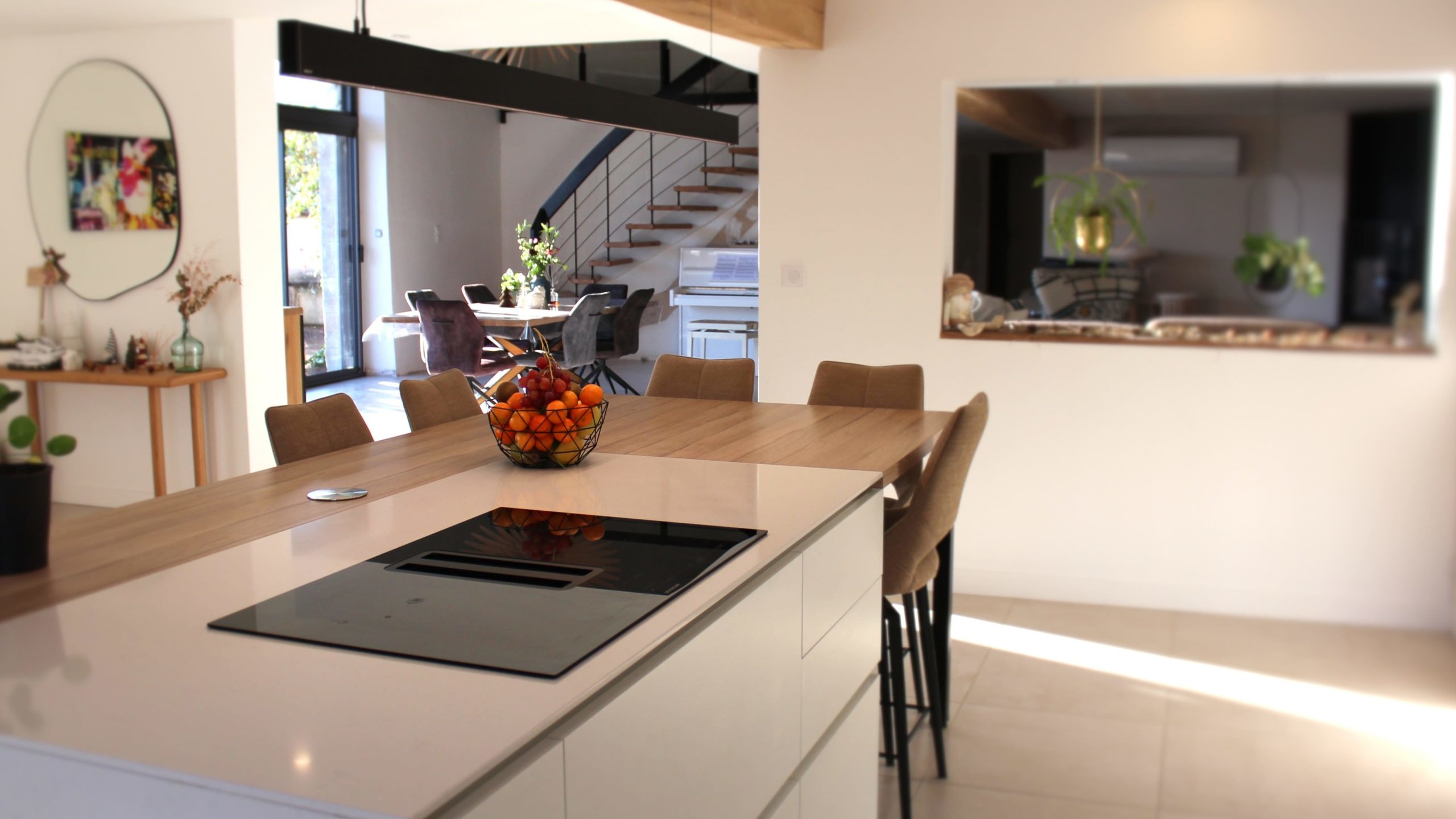 Cette cuisine contemporaine a été pensée comme un véritable espace de vie, alliant esthétique minimaliste, matériaux naturels et fonctionnalité au quotidien. L’association du bois clair et des façades blanches sans poignées apporte une sensation immédiate de luminosité et de sérénité. Au cœur de la pièce, l’îlot central s’impose comme un élément structurant. Il intègre une plaque de cuisson discrète, de nombreux rangements et une grande table en bois massif, idéale pour partager les repas, travailler ou recevoir. Cet aménagement favorise la convivialité tout en conservant une circulation fluide dans l’espace. La poutre en bois apparente traverse la pièce et lui confère un caractère unique, créant un dialogue harmonieux entre architecture traditionnelle et design contemporain. L’éclairage suspendu, aux lignes sobres et graphiques, souligne l’îlot et apporte une touche moderne élégante. Les espaces de rangement intégrés, l’électroménager encastré et les finitions soignées participent à une lecture visuelle épurée. Les touches végétales viennent parfaire l’ensemble en apportant fraîcheur et naturel. Pensée dans les moindres détails, cette cuisine offre un cadre à la fois chaleureux, fonctionnel et durable, parfaitement adapté à un art de vivre moderne.