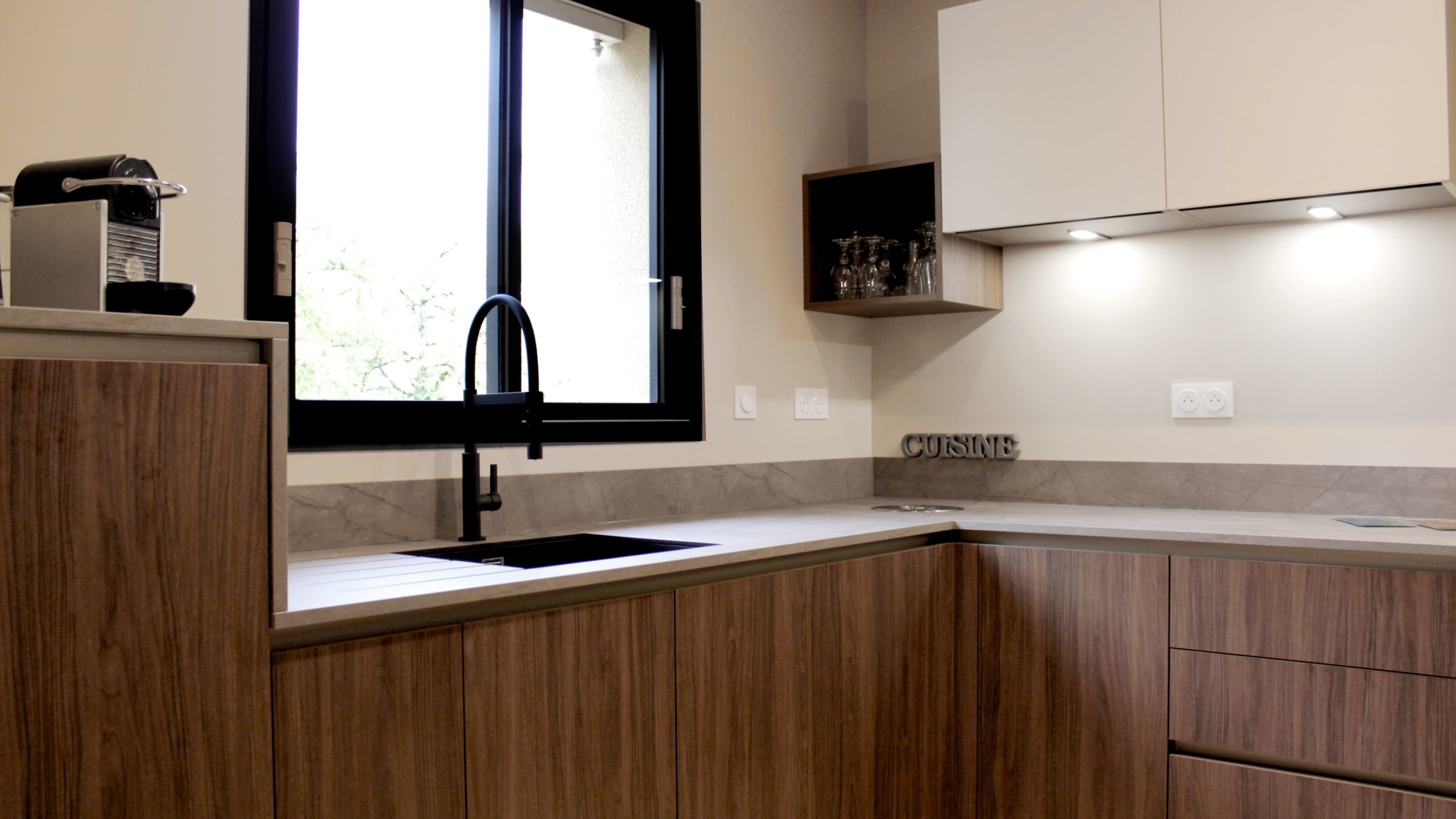 Cuisine moderne en noyer avec meubles clairs, hotte noire, éclairage discret et détails dorés subtils.